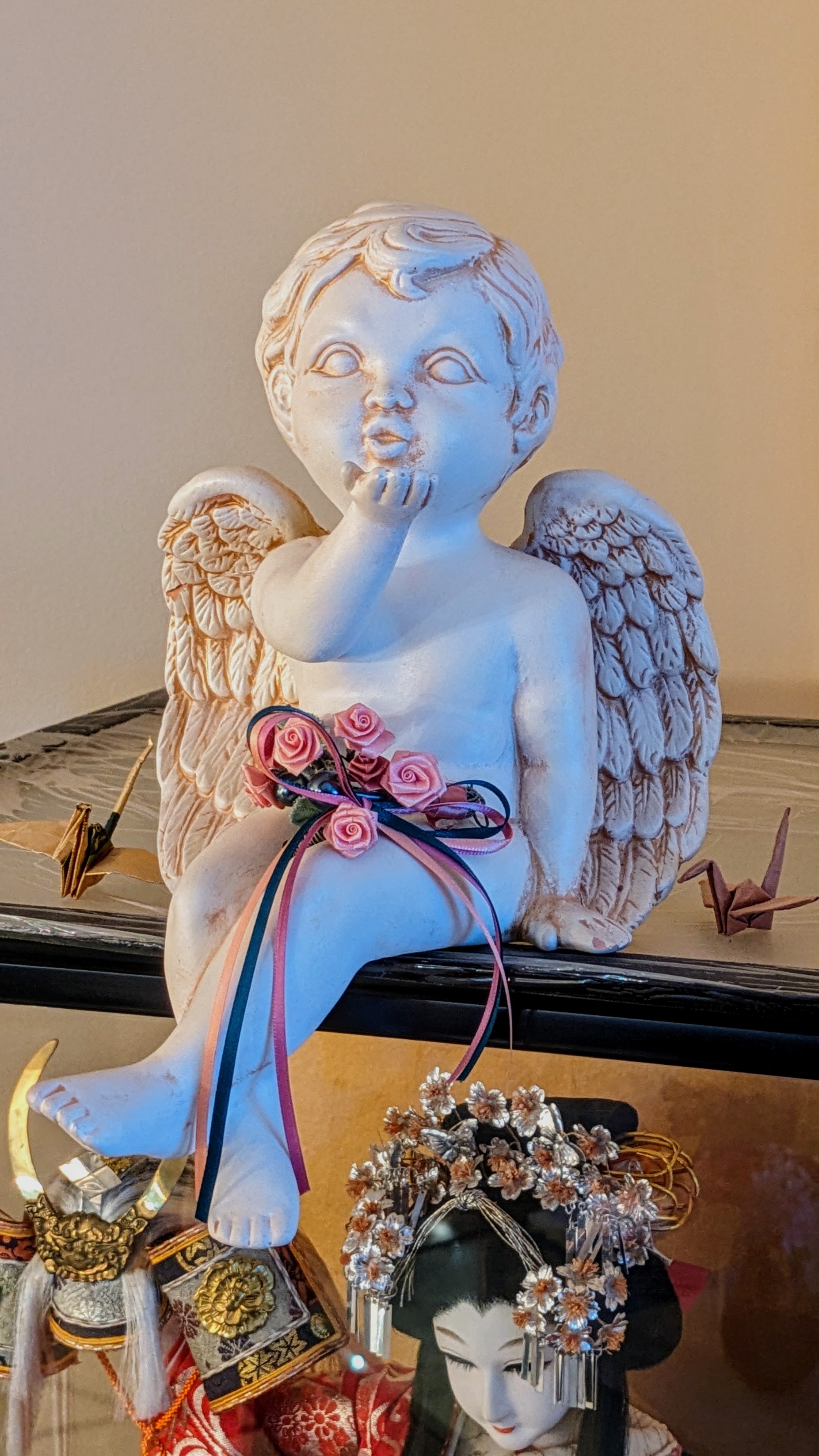 Photo of a seated stone angel blowing a kiss at you with its legs flirtily crossed. Two small origami cranes sit beside it. It is the top layer of the piled photos on this homepage, and must be dragged to reveal the others, unless you are using this alt text. Extended notes: this cherub is found on top of a glass case in my grandparents' house, which contains one of two very fancy Japanese dolls my Obaba brought from Japan to New Jersey in a suitcase in 1969.