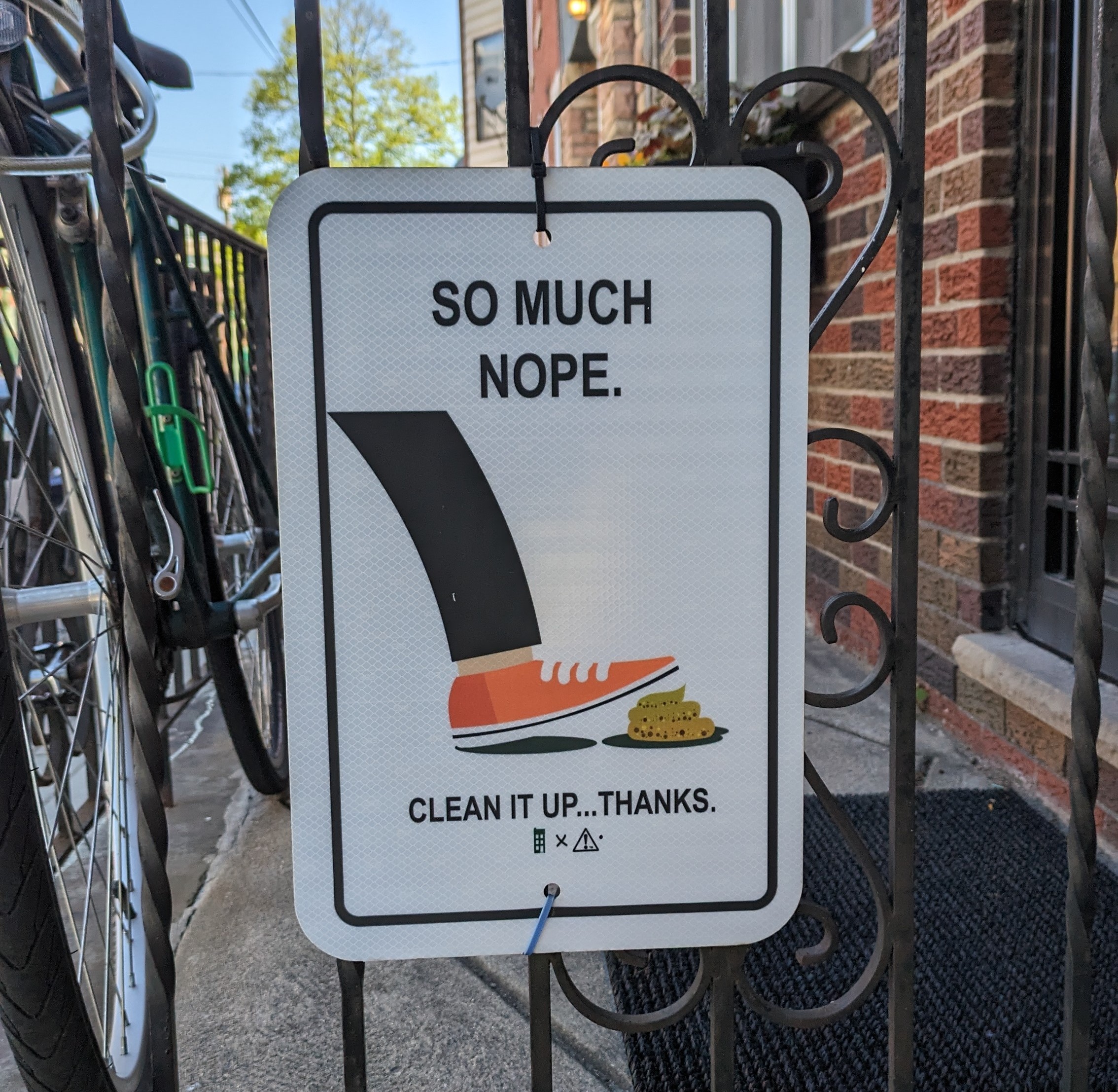 Photograph of a metal sign zip-tied to a porch railing, which depicts a cartoon leg about to step in dog poop. There is very millenial text that reads SO MUCH NOPE, CLEAN IT UP... THANKS