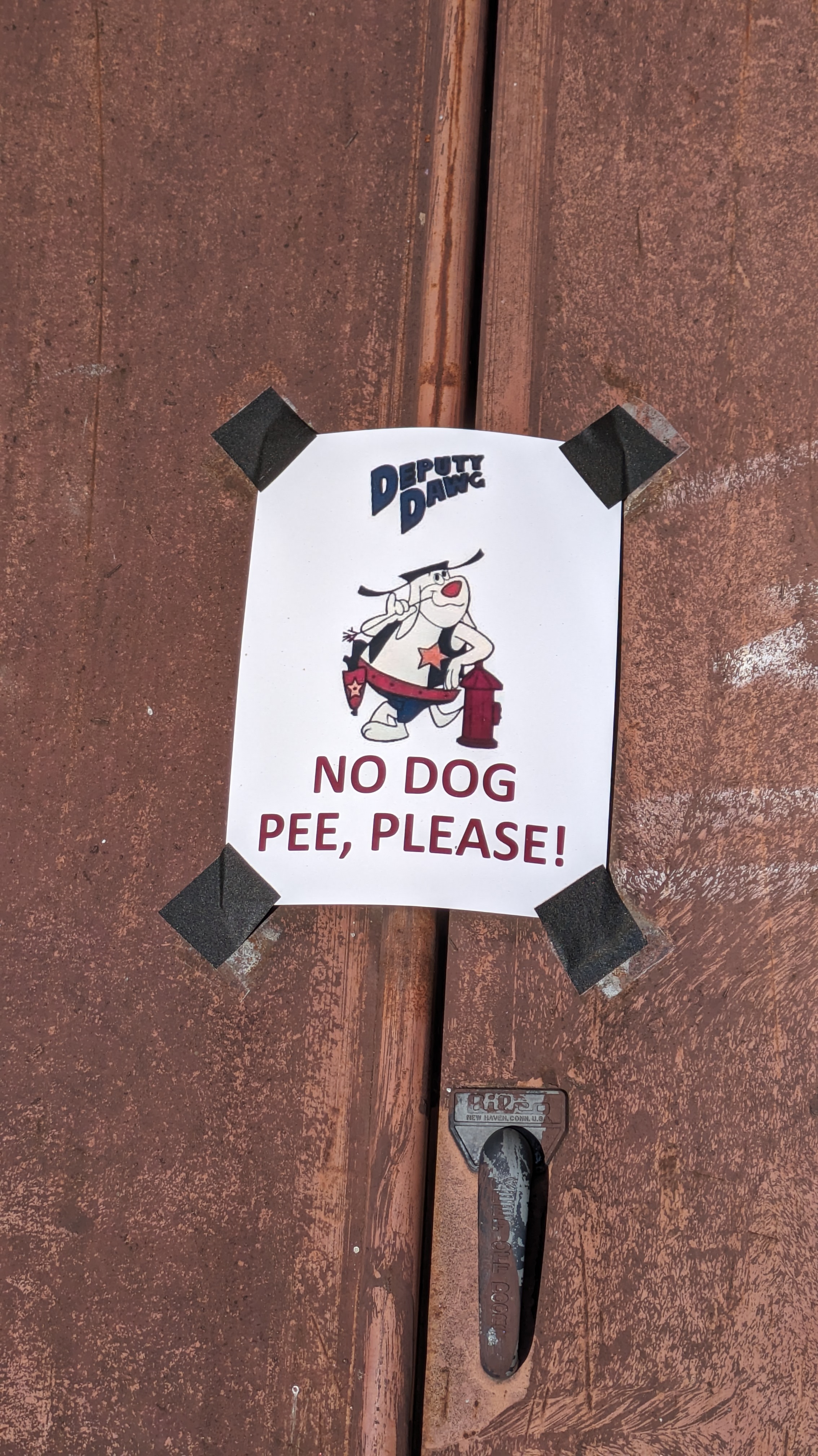 Photograph of a normal letter paper sign duck taped to rusty basement doors. The sign has a cartoon dog labeled Deputy Dawg spelled D. A. W. G. casually leaning on a fire extinguisher and picking his teeth with a piece of straw. He has a gun in a belt holster and a star badge on his vest. Below him reads NO DOG PEE, PLEASE! in all caps red letters.